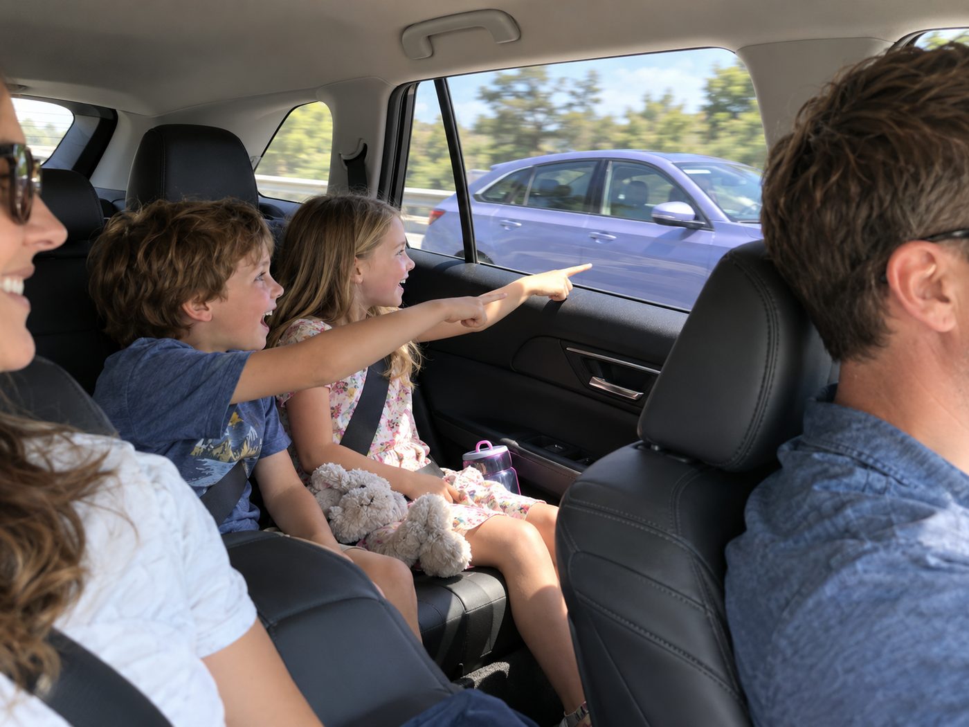 Family on a road trip with kids in the back seat pointing at a periwinkle car alongside